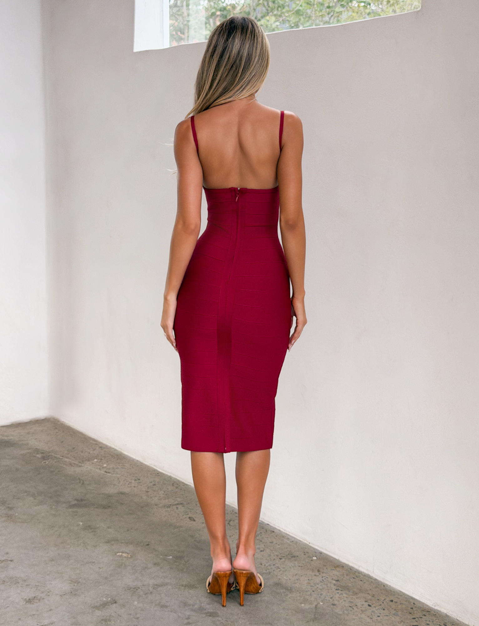 A woman with long blonde hair wears the TIGERMIST MIDORI MIDI BANDAGE DRESS in cherry and tan high heels, standing indoors with her back to the camera near a white wall and window.