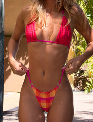 A woman stands outdoors in sunlight and greenery, wearing the TIGERMIST AYANNA REVERSIBLE BIKINI BOTTOM - GINGHAM AND RESORT PLAID, holding its straps. Her face is not visible.
