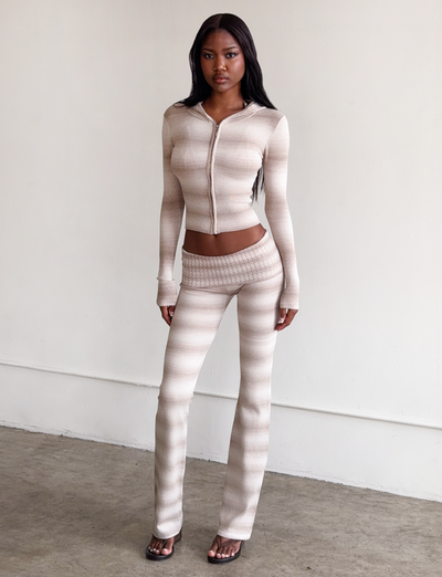 A woman with long dark hair and a neutral expression stands indoors against a white wall, wearing TIGERMIST’s MARISSA FOLDOVER KNIT PANT in Taupe Ombre Stripe with a matching zip-up top and open-toe heels.