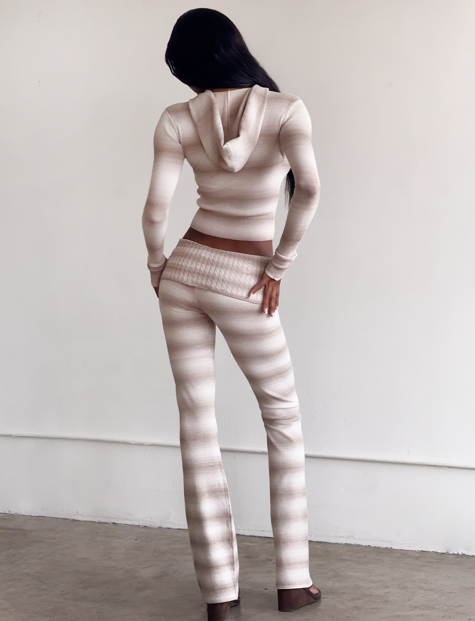 A woman stands with her back to the camera, wearing the TIGERMIST MARISSA JUMPER in taupe ombre stripe with matching flared pants, one hand on her hip in a minimalist indoor setting.