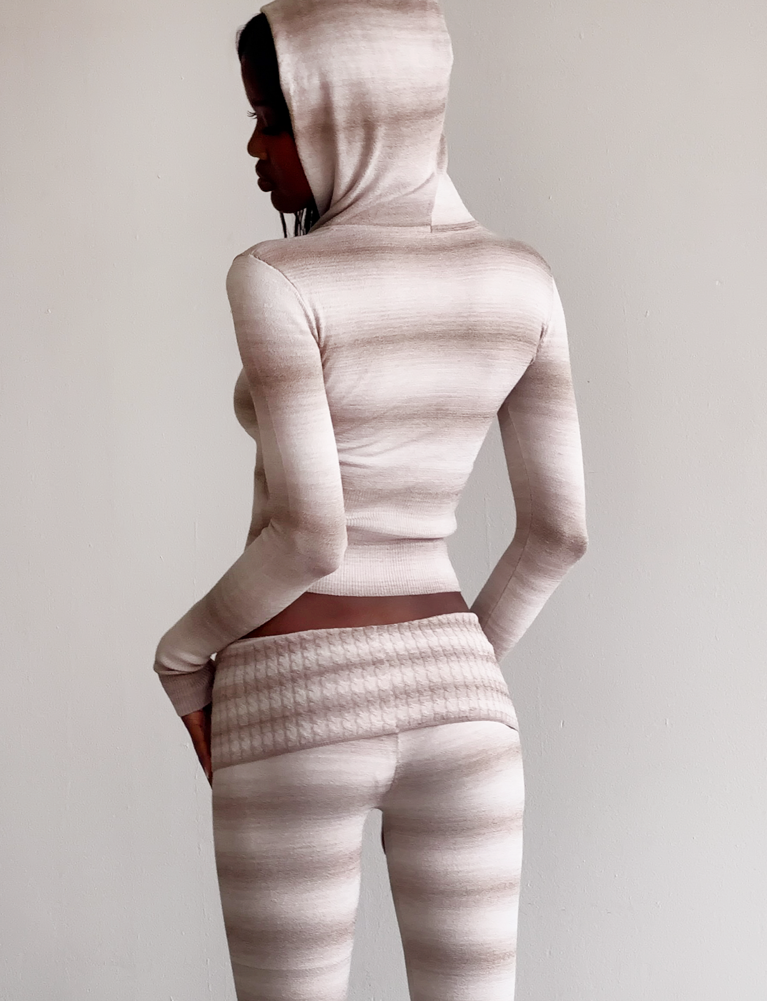 A person stands sideways in the TIGERMIST MARISSA JUMPER - TAUPE OMBRE STRIPE, a fitted, hooded knit jumper with matching high-waisted pants in beige and cream stripes. The background is plain and neutral.