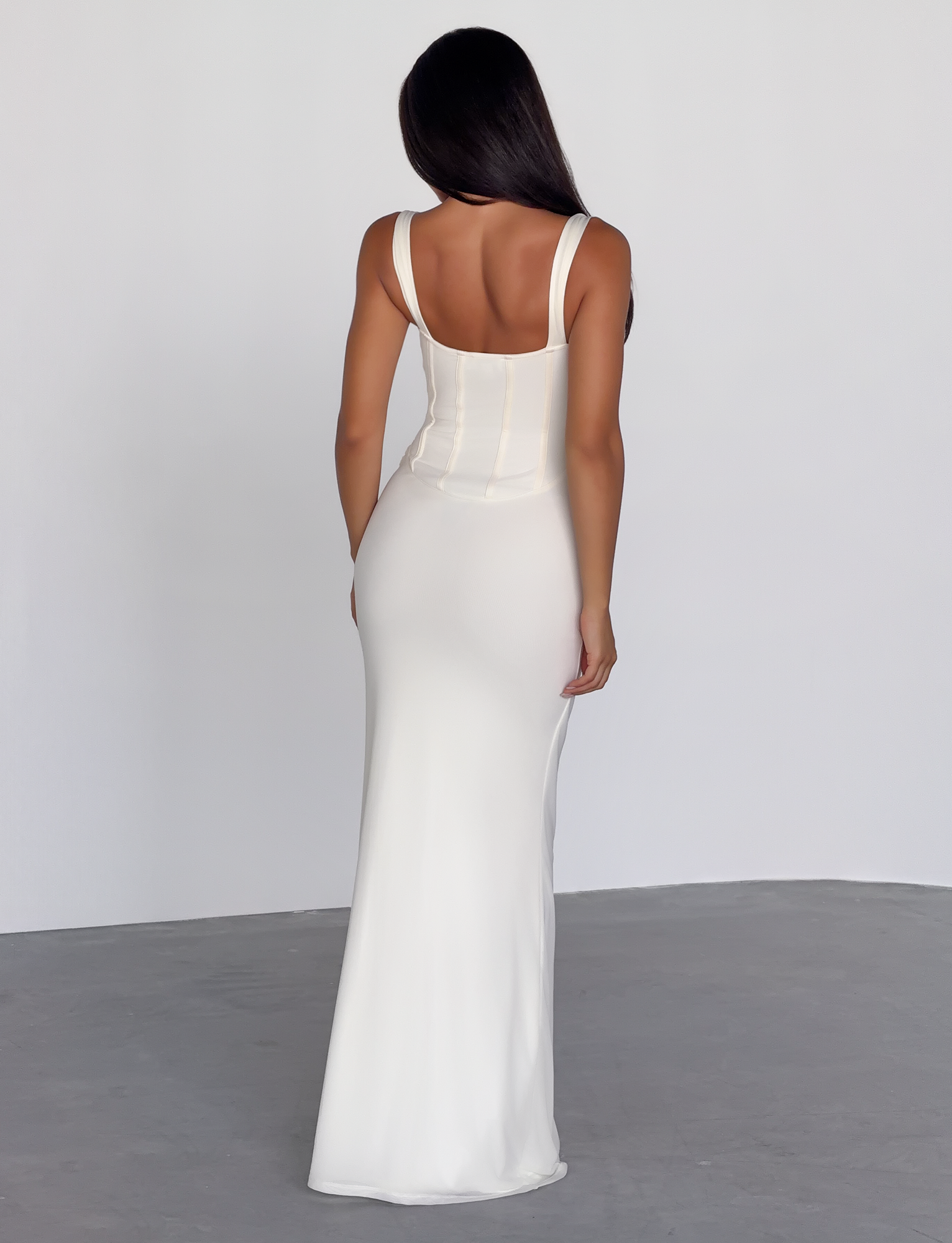 A woman with long dark hair stands facing away, wearing the TIGERMIST GINNY MAXI DRESS in off white—a corseted, form-fitting maxi with ruched bust cups—against a plain, light background.