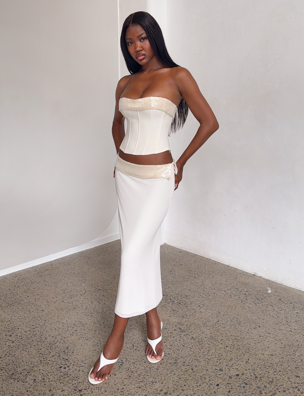 A woman poses indoors wearing the TIGERMIST CHARLYZE MIDI SKIRT in cream with sequin details and a matching strapless corset top, paired with white heeled sandals. She stands on a speckled floor against a plain light wall.