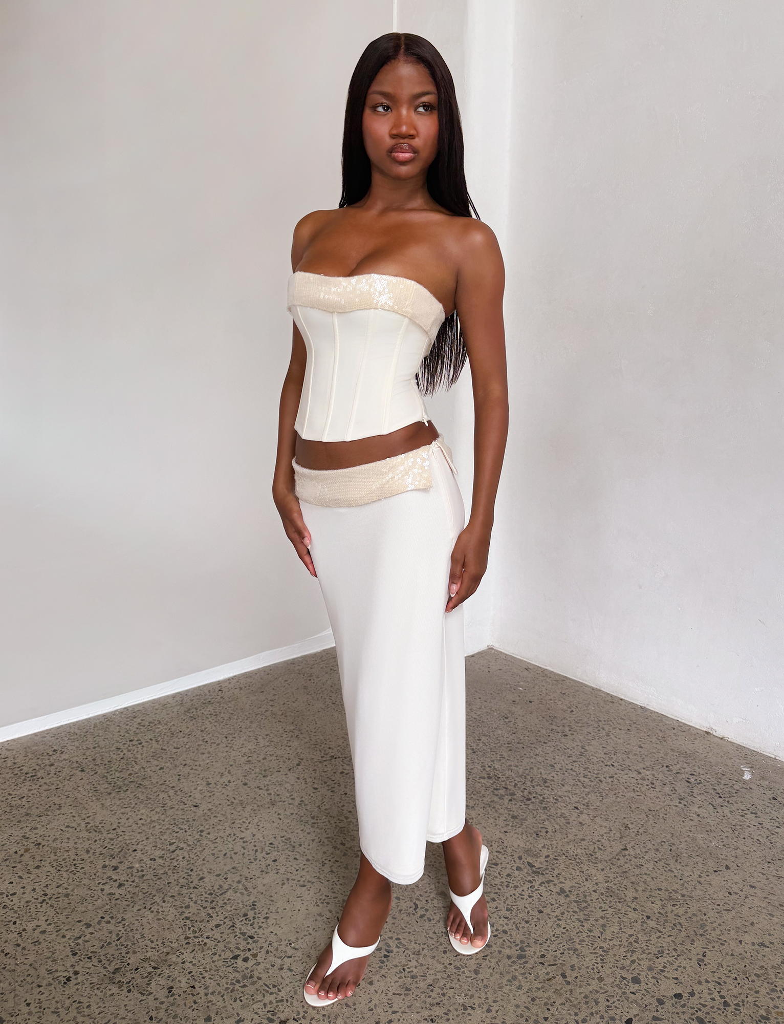 A woman stands indoors wearing the TIGERMIST CHARLYZE MIDI SKIRT in cream, paired with a strapless corset top with gold accents, white heeled sandals, and has long straight hair. The setting features white walls and a concrete floor.