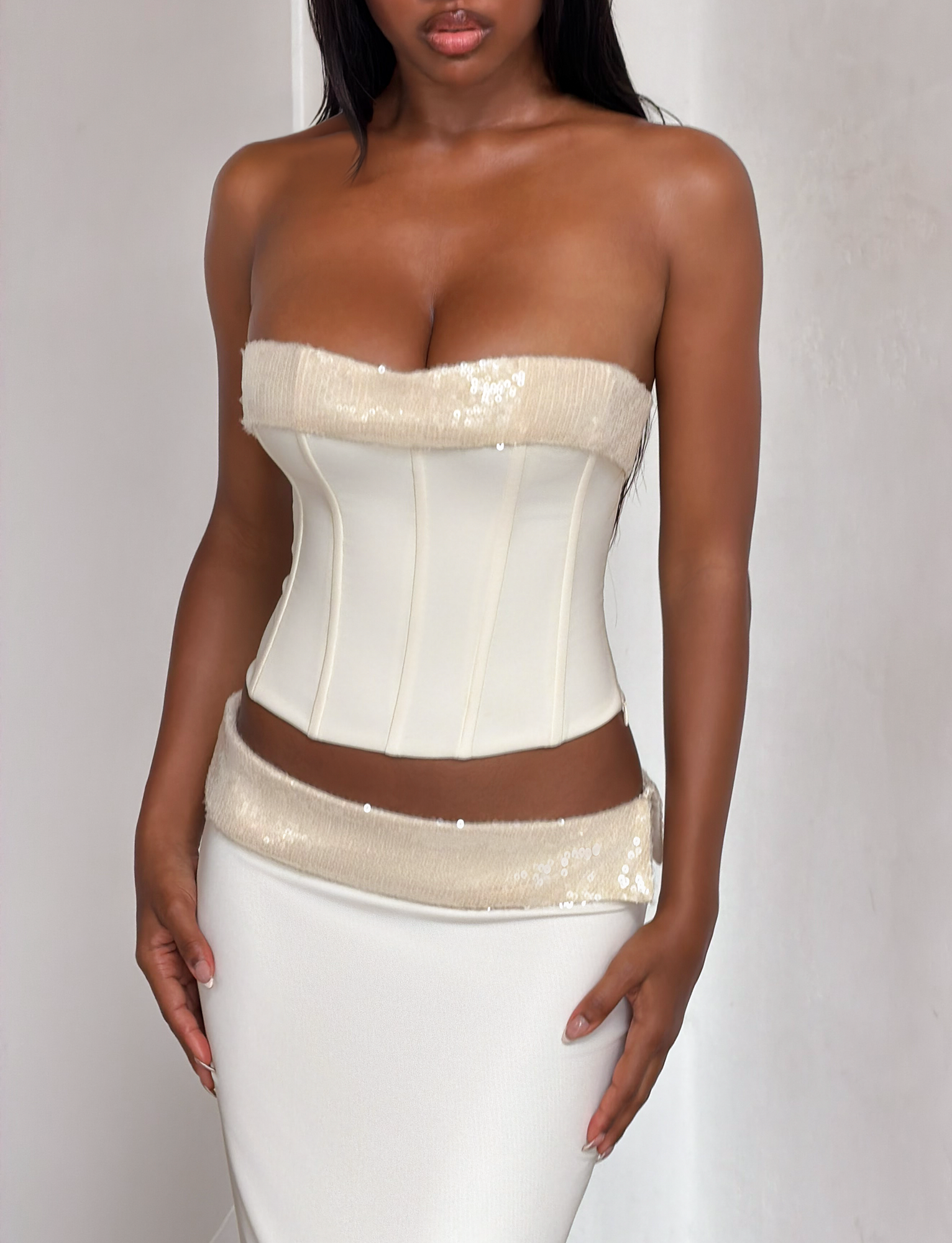 A woman wears the TIGERMIST CHARLYZE BUSTIER in cream, a white strapless top with sequin trim, lace-up back, and matching high-waisted skirt. The elegant, form-fitting set is photographed against a plain light background.