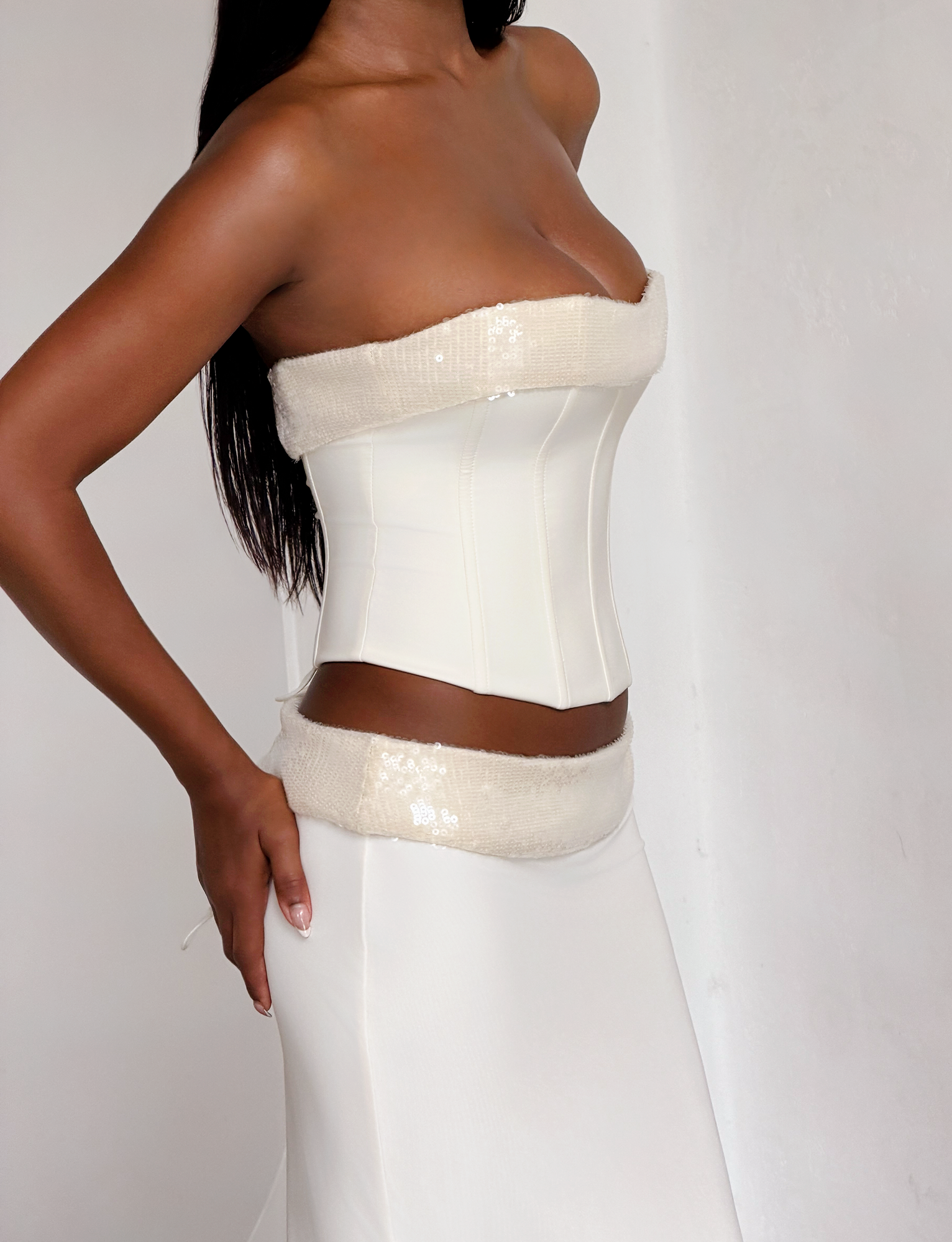 A woman poses in a minimalist space wearing the TIGERMIST CHARLYZE BUSTIER in cream with a matching skirt, featuring a subtle sheen, textured trim, and chic lace-up back, her hand resting on her hip.
