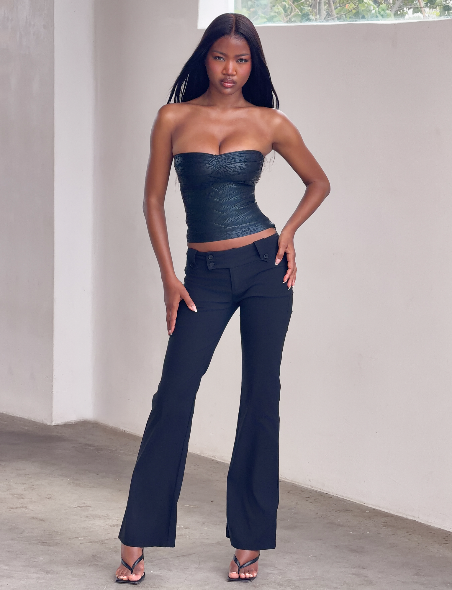 A woman poses confidently indoors against a white wall, wearing the TIGERMIST NIYA METALLIC BANDAGE TOP in black with black pants and heeled sandals. She has long, straight hair and rests one hand on her hip.
