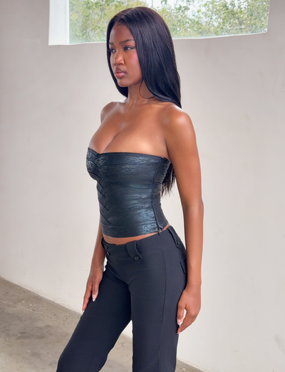 A woman with long straight hair wears the TIGERMIST NIYA METALLIC BANDAGE TOP in black and black pants, standing indoors by a white wall with a window, looking to the side.