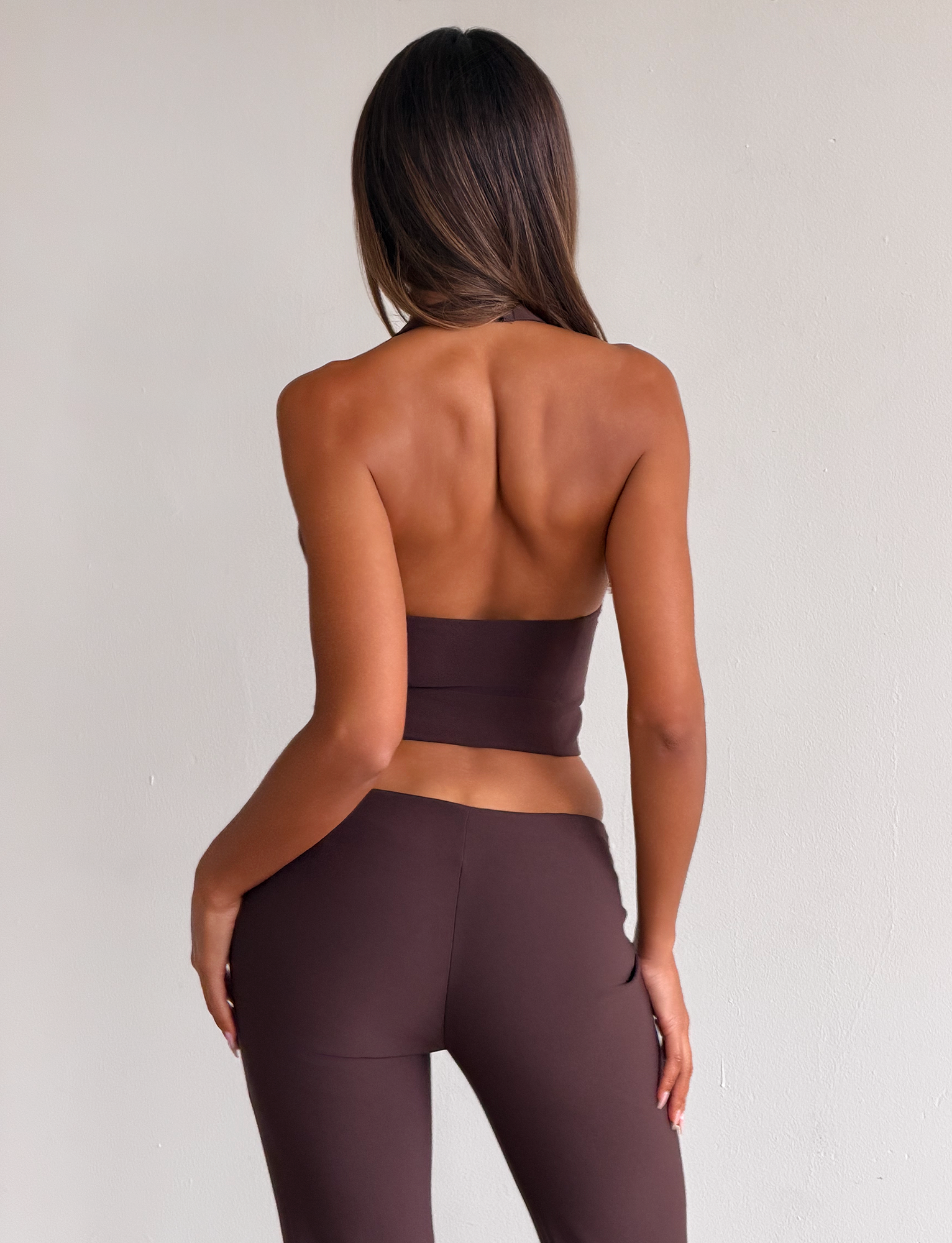 A woman with long, straight hair faces away from the camera, wearing the TIGERMIST KITTIE BUCKLE TOP - CHOCOLATE with matching brown pants against a plain light wall.