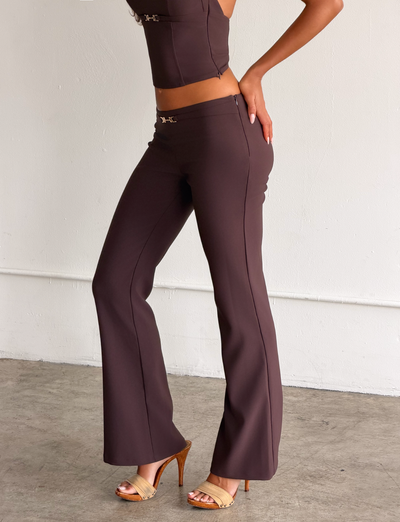 A woman wears the TIGERMIST KITTIE BUCKLE PANT in chocolate—high-waisted, flared, and made from stretch suiting fabric—with a matching spaghetti strap top and high-heeled sandals, standing on a concrete floor against a plain white wall.