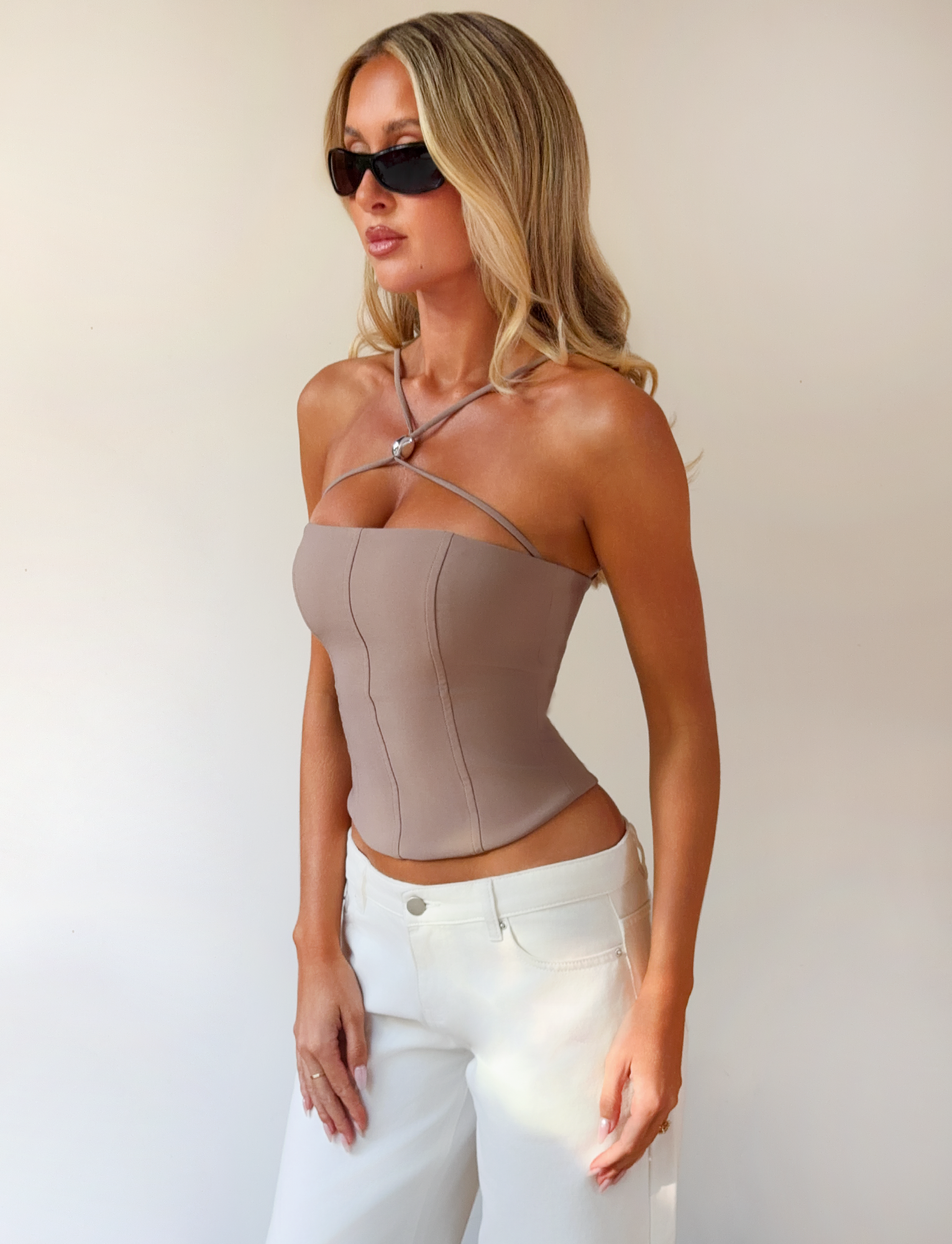 A woman with long blonde hair and black sunglasses wears the TIGERMIST TAMORA CORSET TOP in taupe with adjustable straps and white pants, standing against a plain light background.