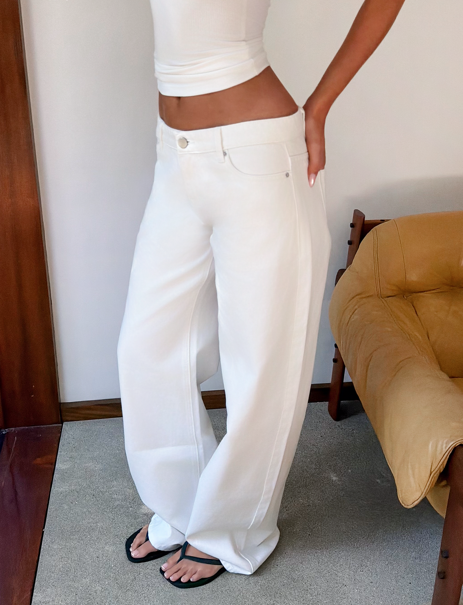 A person stands indoors by a tan chair, wearing TIGERMIST MALIYA JEAN - WHITE with a low rise and extra long inseam, a fitted white tank top, and black sandals, one hand on their hip. Their head is not visible.