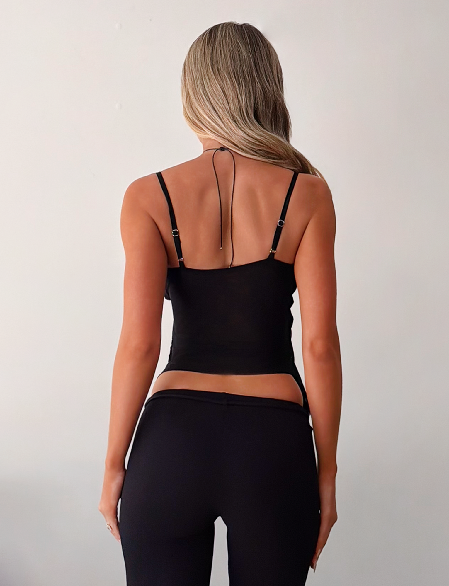A woman with long, wavy blonde hair stands with her back to the camera, wearing the TIGERMIST ALDEANA TOP in black and black pants against a plain, light background.