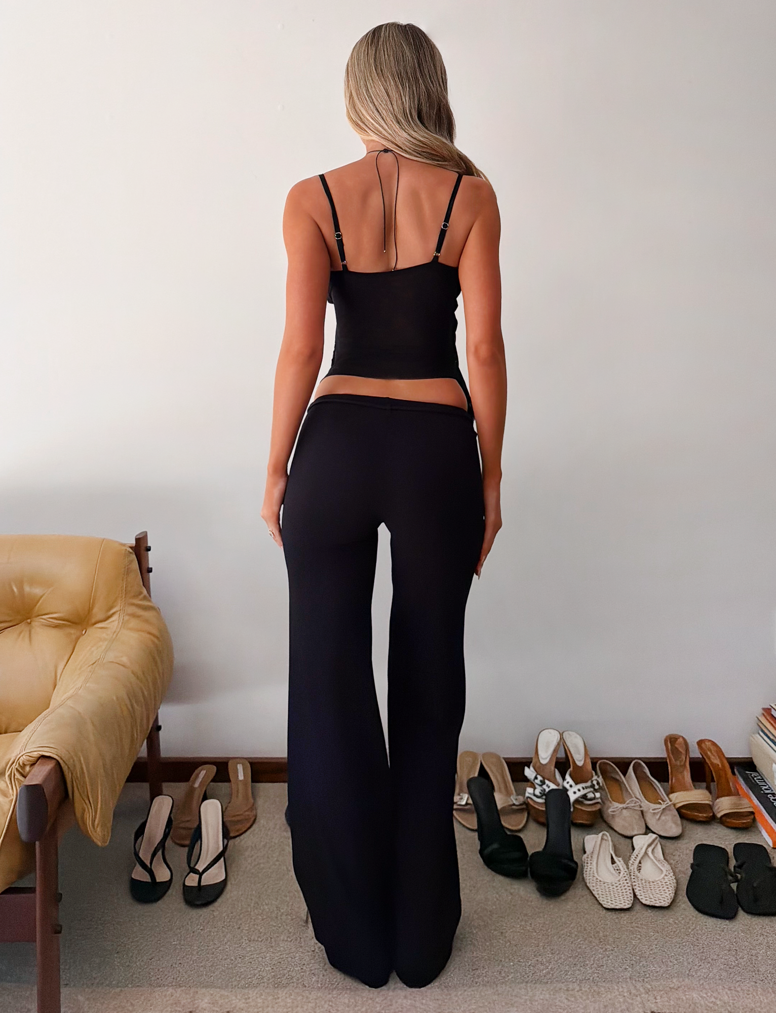 A woman with long blonde hair stands with her back to the camera, wearing the TIGERMIST KAILI PANT in black and a black camisole. Various shoes are lined up behind her, and a beige chair is on the left.