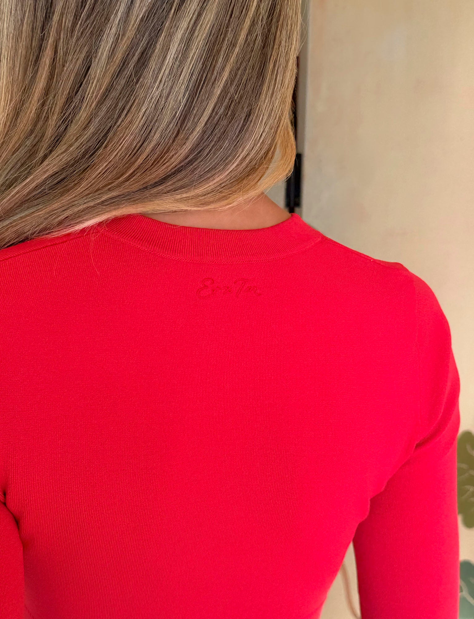 A person with long, light brown hair is shown from the back, wearing the TIGERMIST DAMARIS CARDIGAN in red, a bright button-up knit featuring subtle text near the neckline.