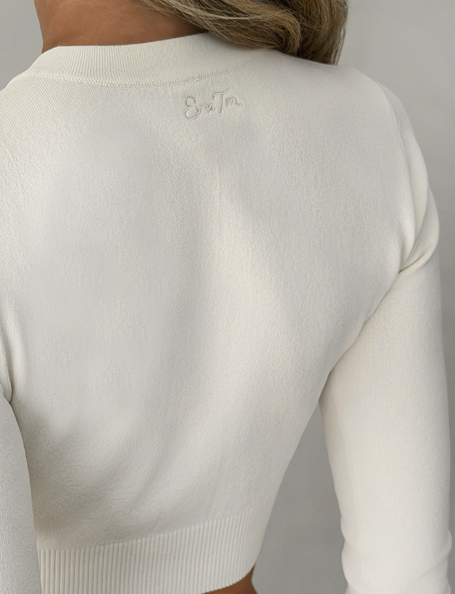 A close-up of a person wearing the TIGERMIST DAMARIS CARDIGAN in cream, a fitted, long-sleeve knit with subtle embroidered text near the neckline. The cropped silhouette highlights the back and shoulders, with hair partially visible.