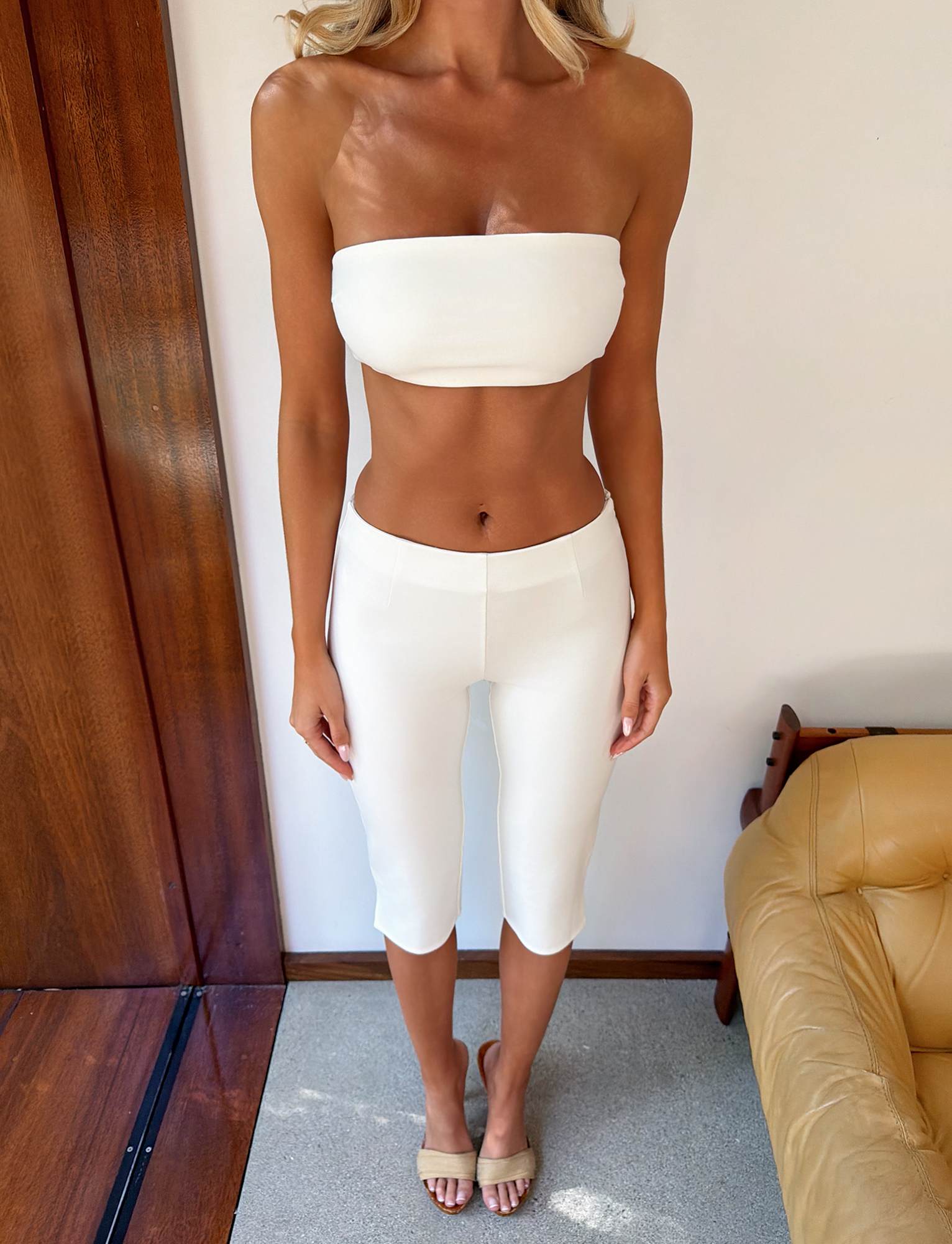 A woman stands indoors wearing the TIGERMIST PILAR BANDEAU in white, paired with white fitted capri pants and beige sandals. She is near a wooden wall and a tan leather chair on a carpeted floor.