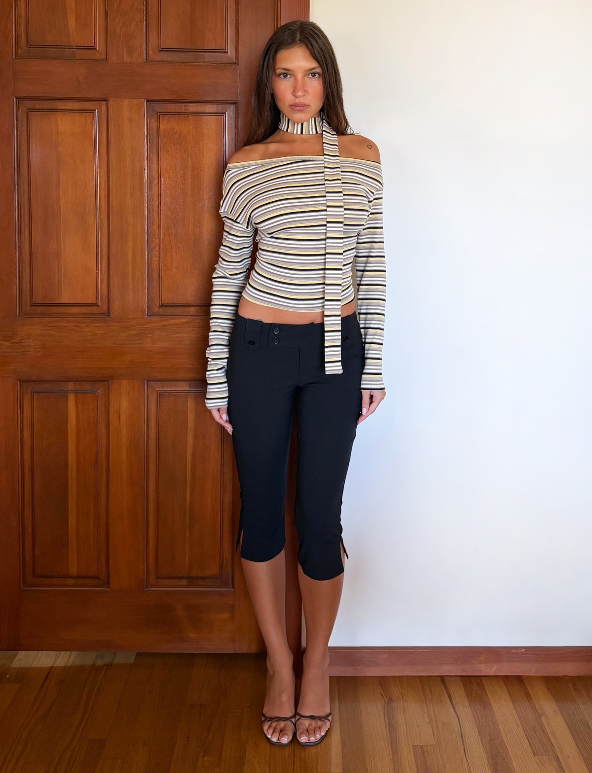 A woman stands indoors against a wooden door, wearing the TIGERMIST KIERSTEN SCARF TOP - MULTISTRIPE with an off-shoulder neckline and long tie, paired with black cropped pants, clear-strap sandals, and her long straight hair down.