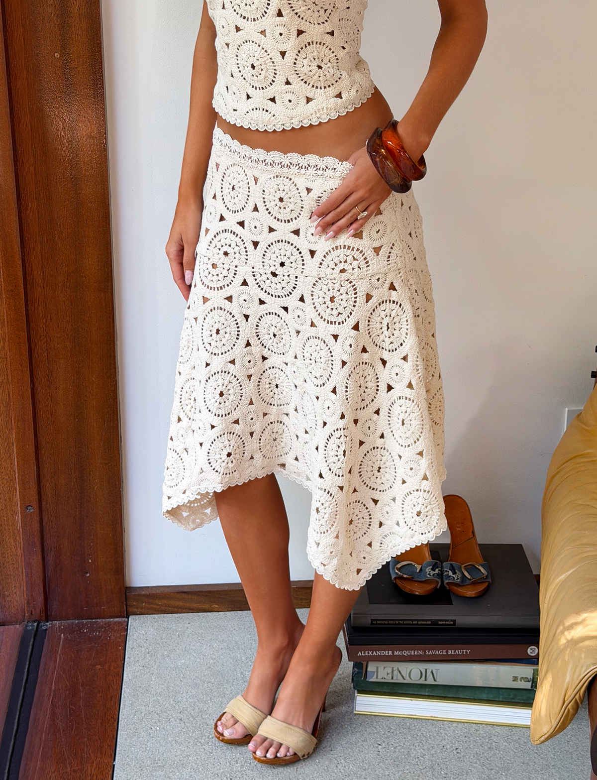 A person stands indoors wearing the TIGERMIST SYNTHIA CROCHET MIDI SKIRT in cream with a matching top, tan sandals, and chunky brown bracelets. Their hand is on their hip; nearby are a stack of books and a gold cushion.