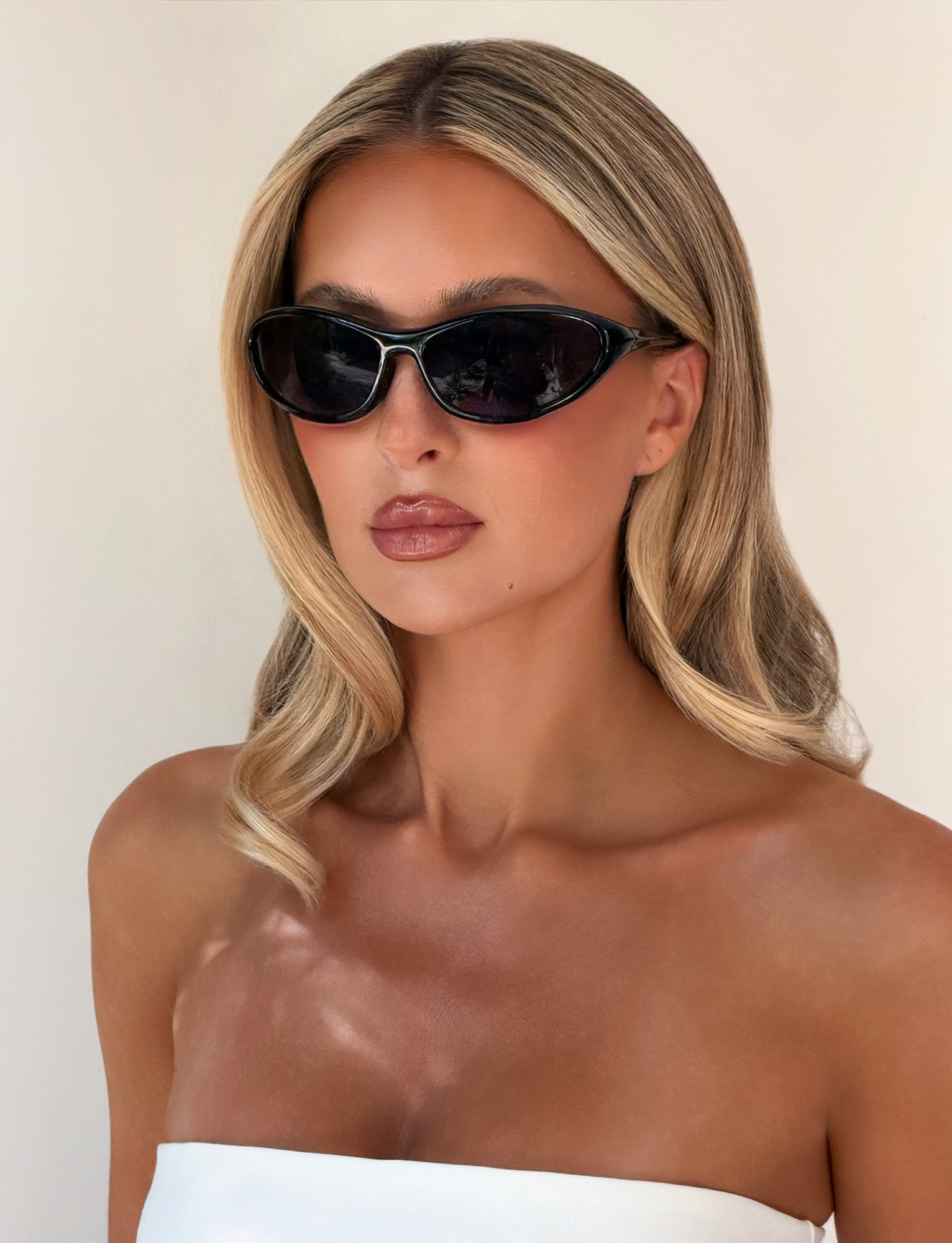 A woman with long, blonde hair and tanned skin wears TIGERMIST RIVAH FASHION GLASSES - BLACK, featuring reflective black lenses, paired with a white strapless top against a plain light background.