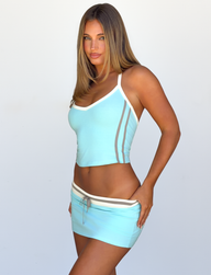 A woman with long hair stands against a plain background, looking at the camera with a neutral expression while wearing the TIGERMIST ALYSIA TOP in Aqua and a matching mini skirt.