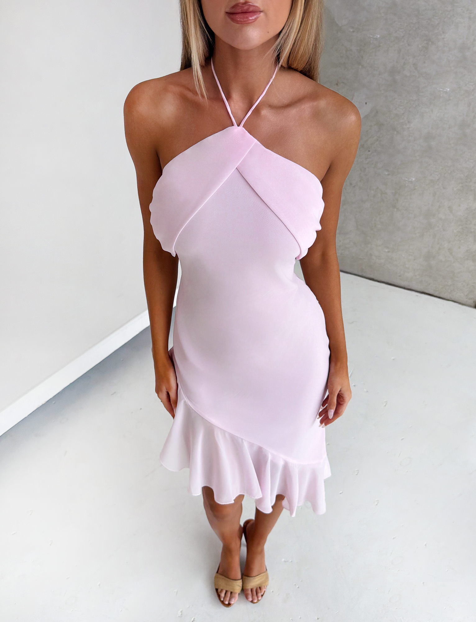 A woman stands indoors on a white floor against a neutral background, wearing the TIGERMIST ELARINA MIDI DRESS in pink with ruffles, a halter-neck, and an asymmetrical hemline. Her face is partially out of frame.