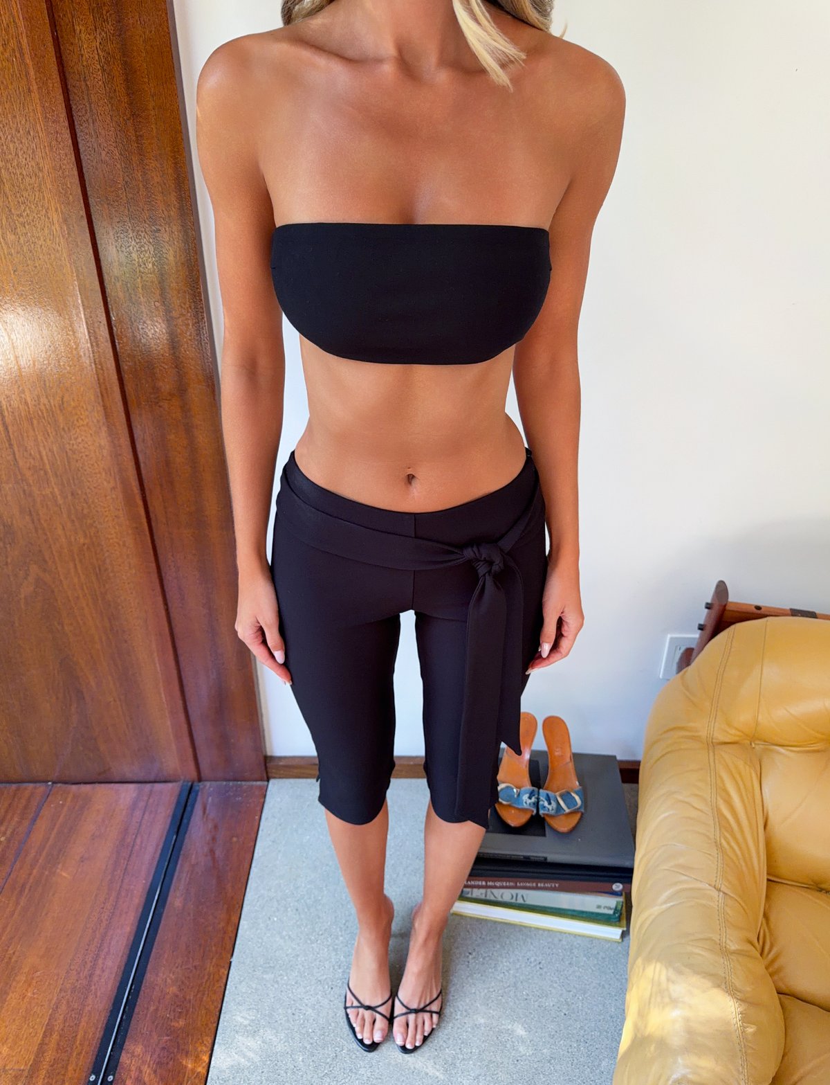 A woman is shown from the shoulders down wearing the TIGERMIST PILAR BANDEAU - BLACK indoors, paired with cropped black pants and black sandals. A yellow chair and a stack of books topped with sandals are beside her.
