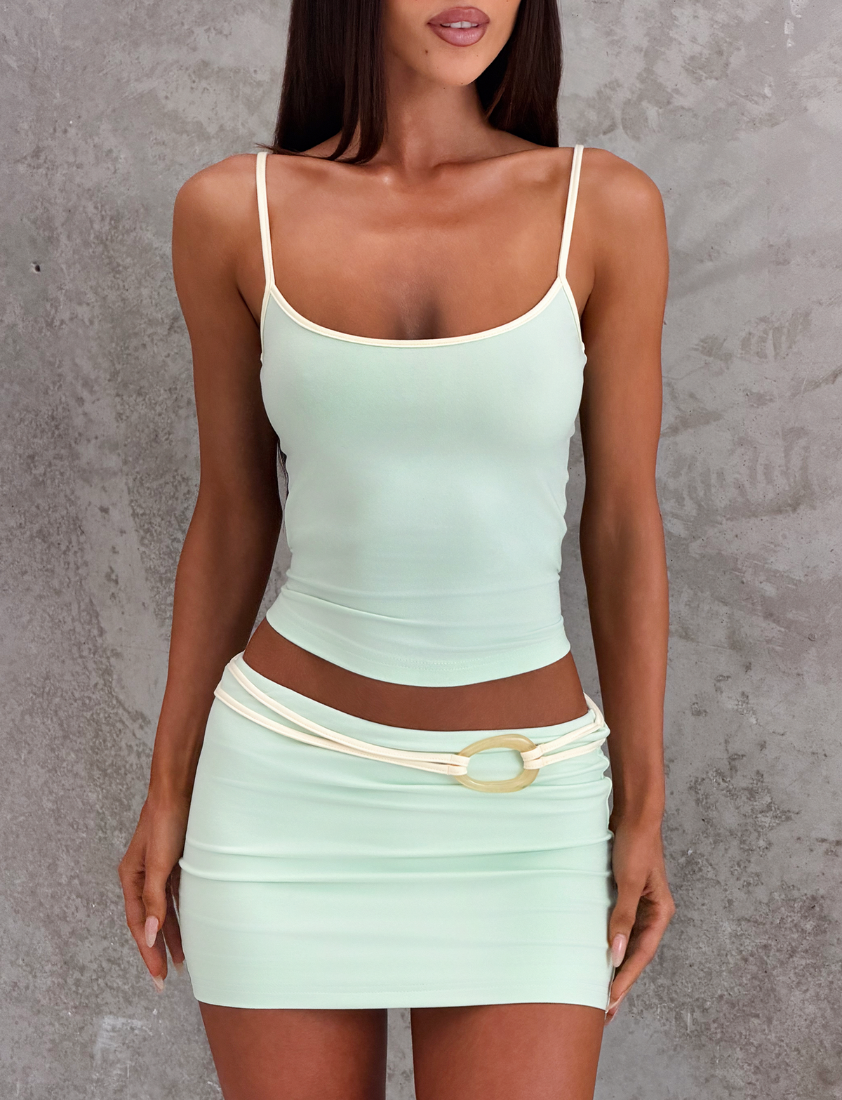 A woman stands against a textured gray wall, wearing the TIGERMIST GREER TOP in mint green with adjustable straps and a scoop neckline, paired with a matching mini skirt featuring a decorative belt and gold oval buckle. Her face is partially out of frame.