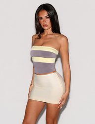A woman with long, straight dark hair and a neutral expression stands against a white background, wearing the TIGERMIST EOS TOP in grey and yellow—a colour blocked, strapless piece featuring pastel stripes.