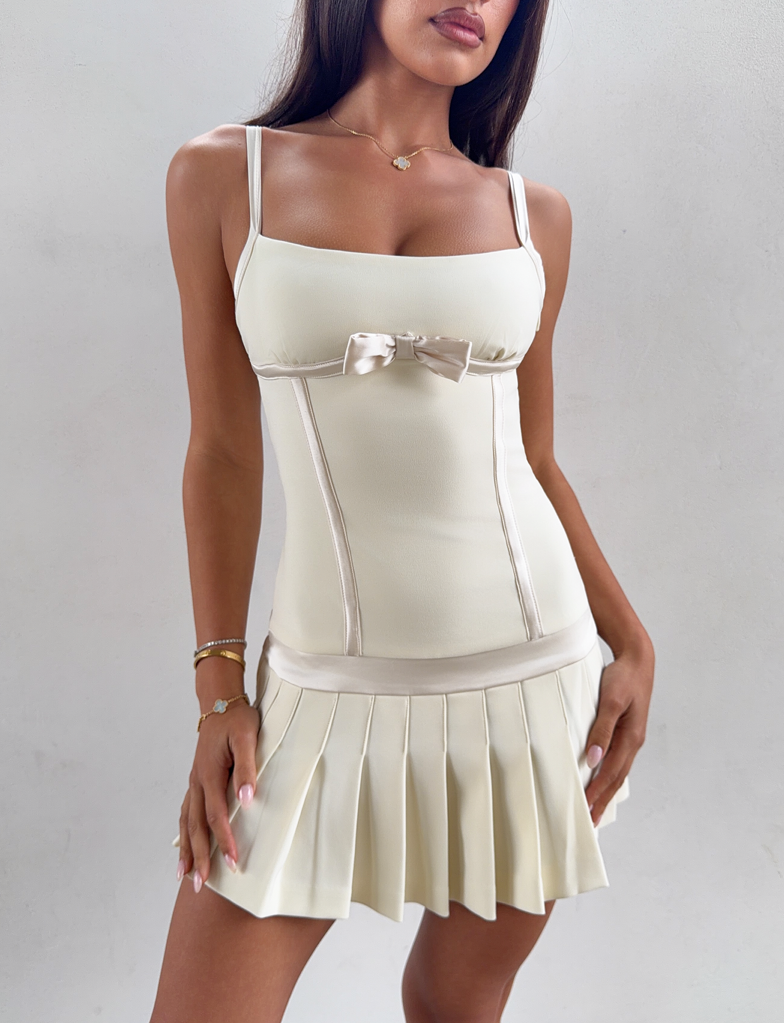 A woman stands against a plain light background wearing the TIGERMIST MARLOW MINI DRESS in cream. This sleeveless dress features adjustable shoulder straps, a small bow accent at the chest, and delicate pleats for an effortlessly elegant look.