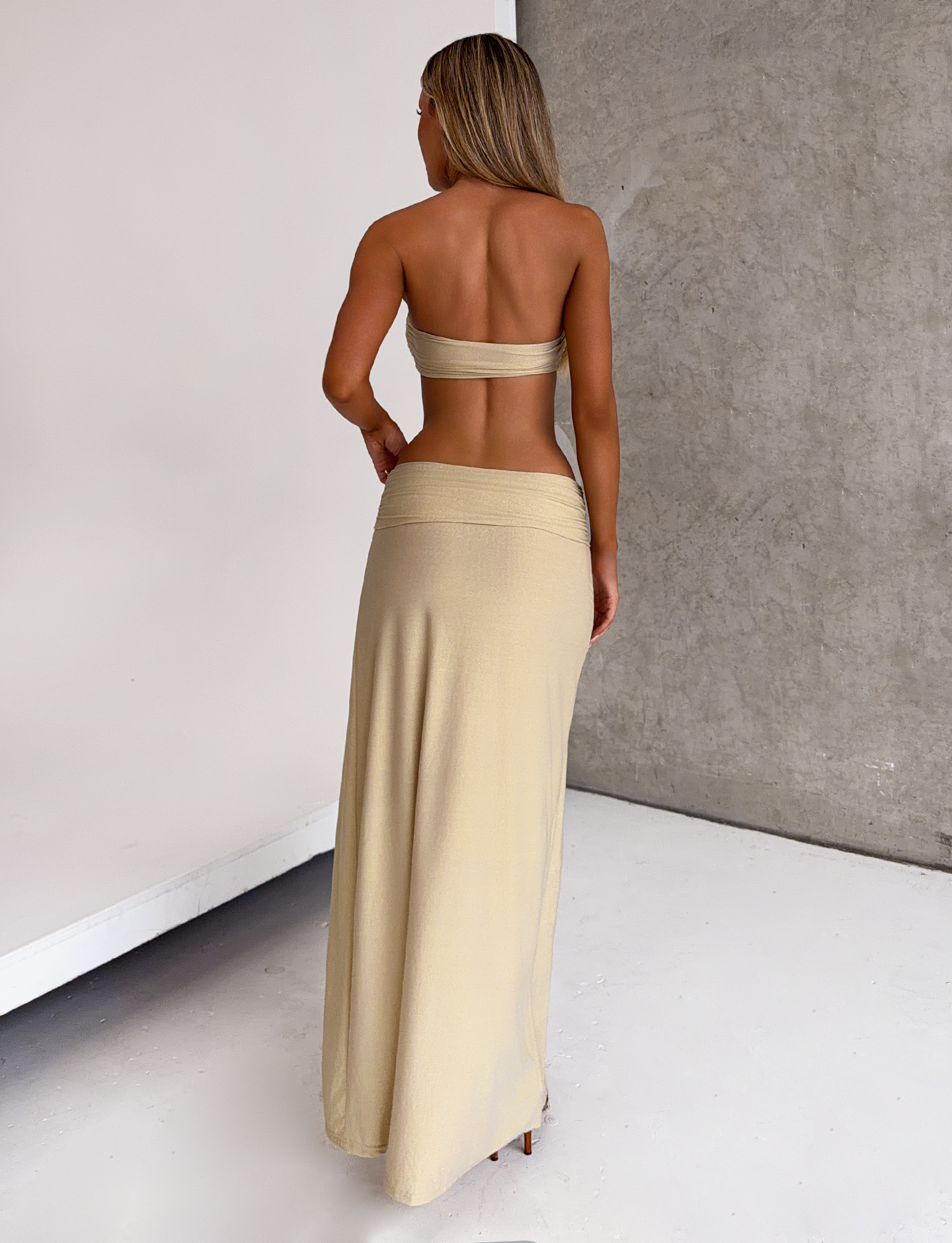 A woman faces away from the camera in a gold TIGERMIST KYNLEE 2.0 MAXI DRESS with a sweetheart neckline, set against a plain light floor and gray textured wall.