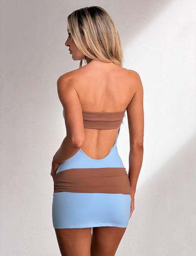 A woman with long blonde hair stands sideways against a white textured wall, wearing the TIGERMIST EOS MINI DRESS in soft blue and mocha featuring a strapless, color-blocked design with an open back and ruched detailing.