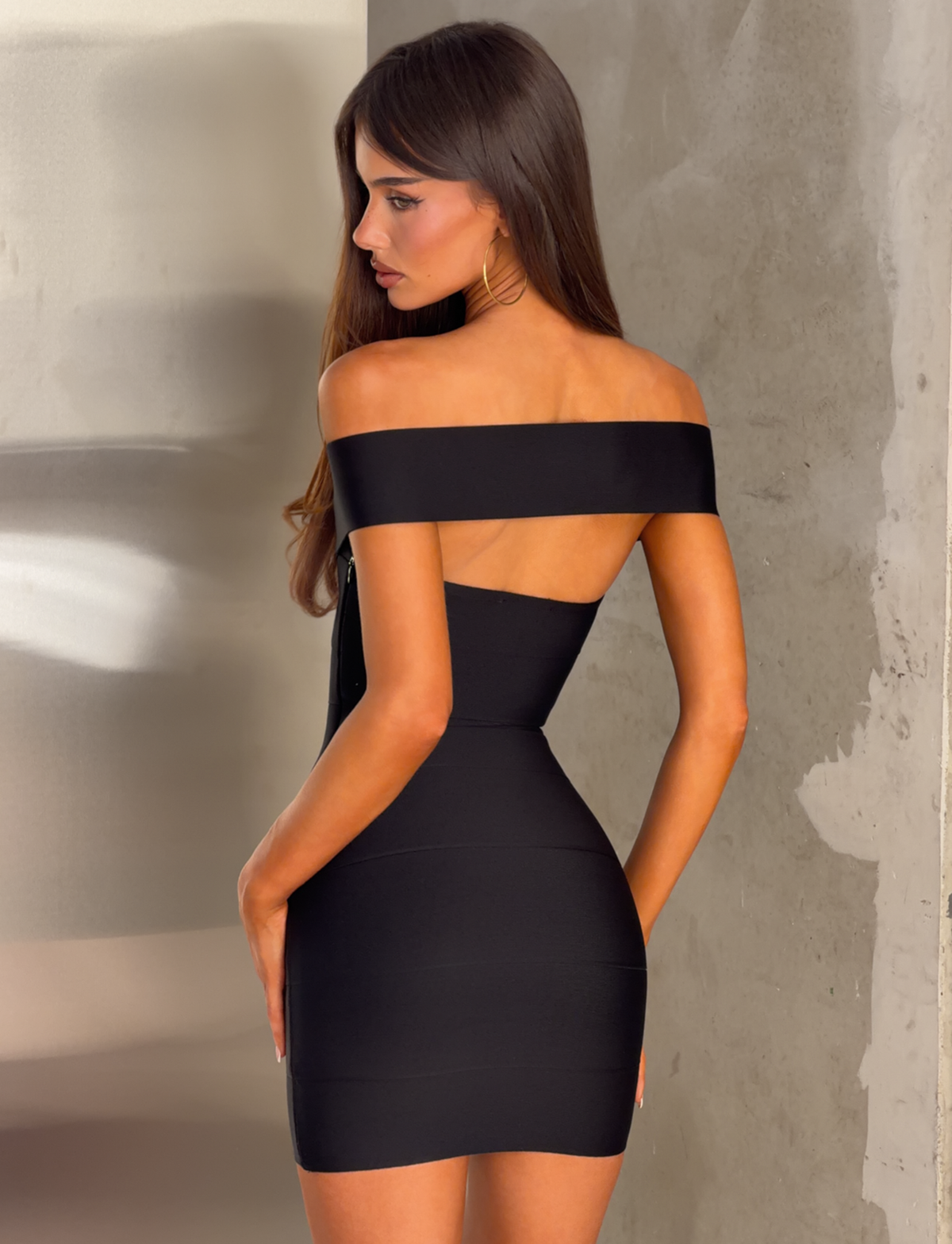 A woman with long brown hair is seen standing sideways and looking over her shoulder against a neutral, modern background, wearing the TIGERMIST ANTONEA BANDAGE MINI DRESS in black, which features an off-shoulder design and a flattering sweetheart neckline.
