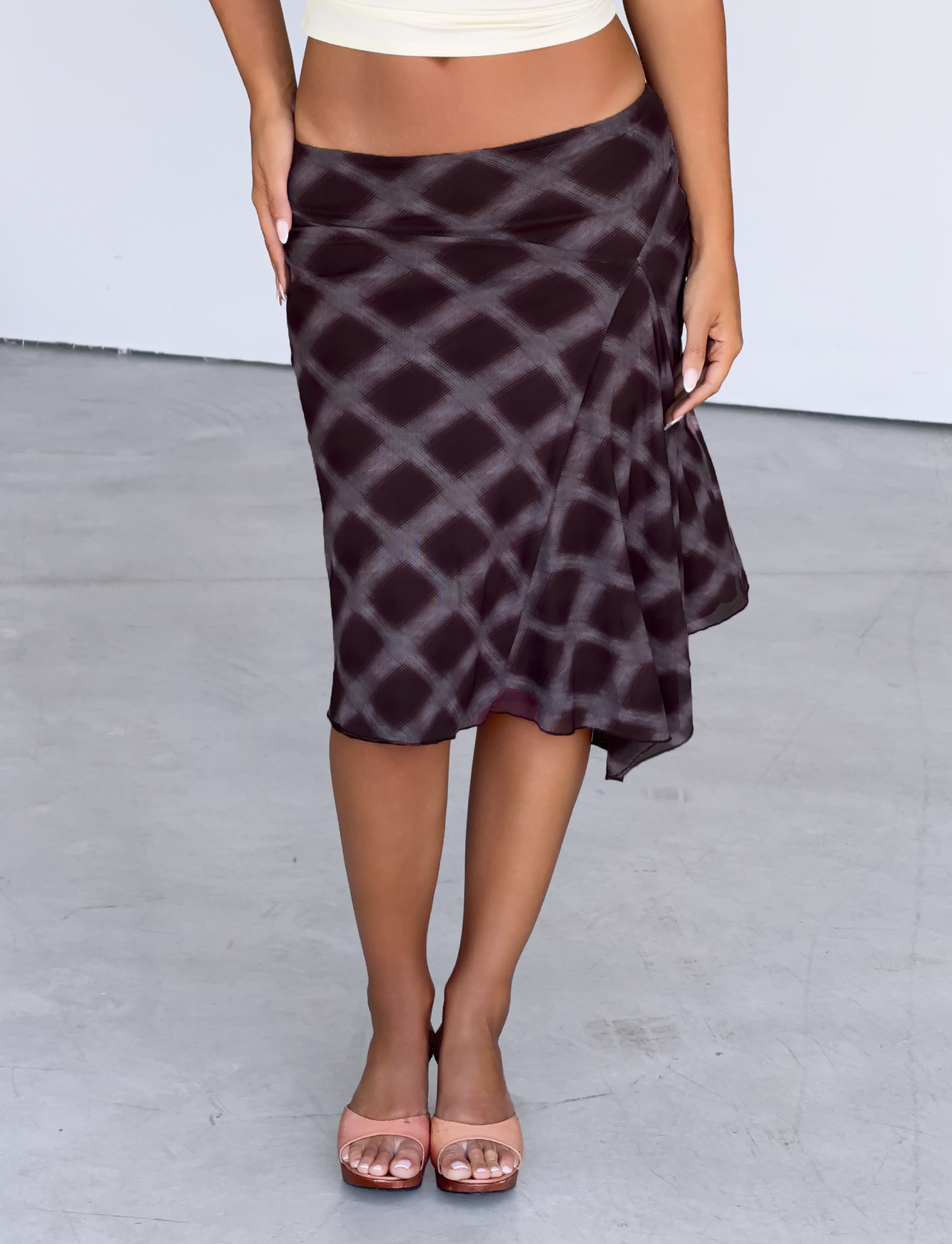 A person stands on a smooth, light gray floor wearing the TIGERMIST SAFFRON MIDI SKIRT in mahogany check and light pink open-toed heels. The upper body and face are not visible.
