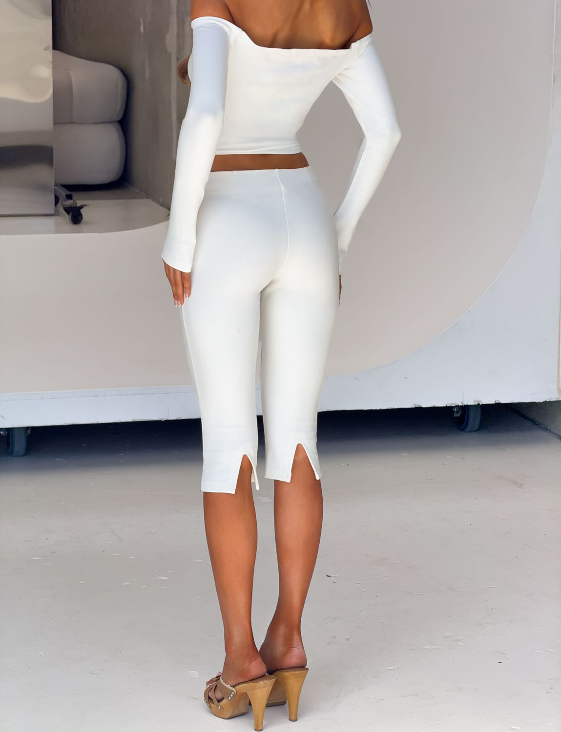 A woman stands facing away, wearing the TIGERMIST IDINA CAPRI in ivory—sleek low-rise capri pants made from ponte fabric with subtle slits at the calves. She styles them with a white off-shoulder crop top and tan heeled sandals, set against a minimal and modern background.