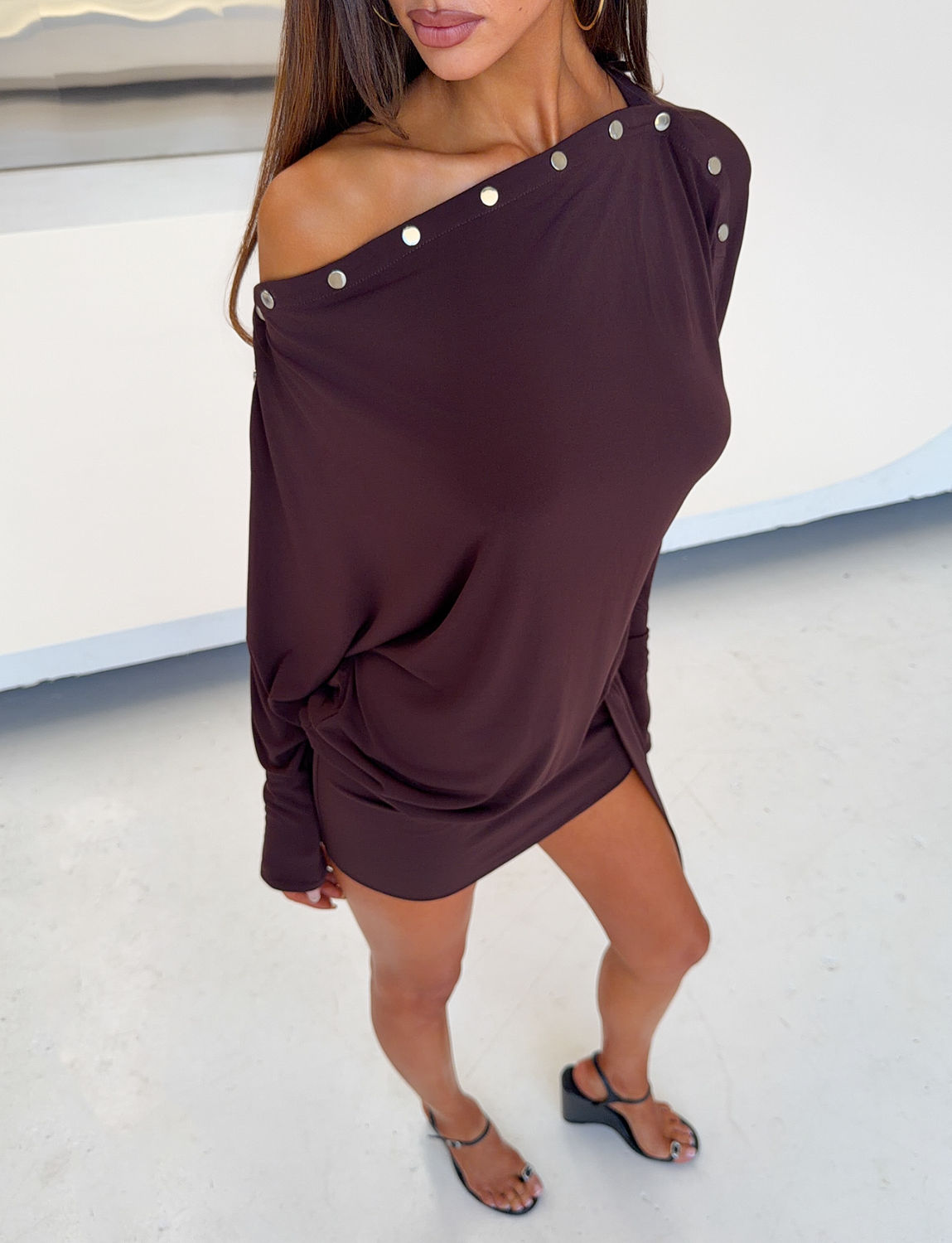 A woman is seen from the shoulders down standing indoors on a light floor, one leg slightly forward, wearing the TIGERMIST RAVENNA LONG SLEEVE MINI DRESS in Dark Chocolate—a loose boat neck style adorned with metallic studs along the neckline—paired with black sandals.