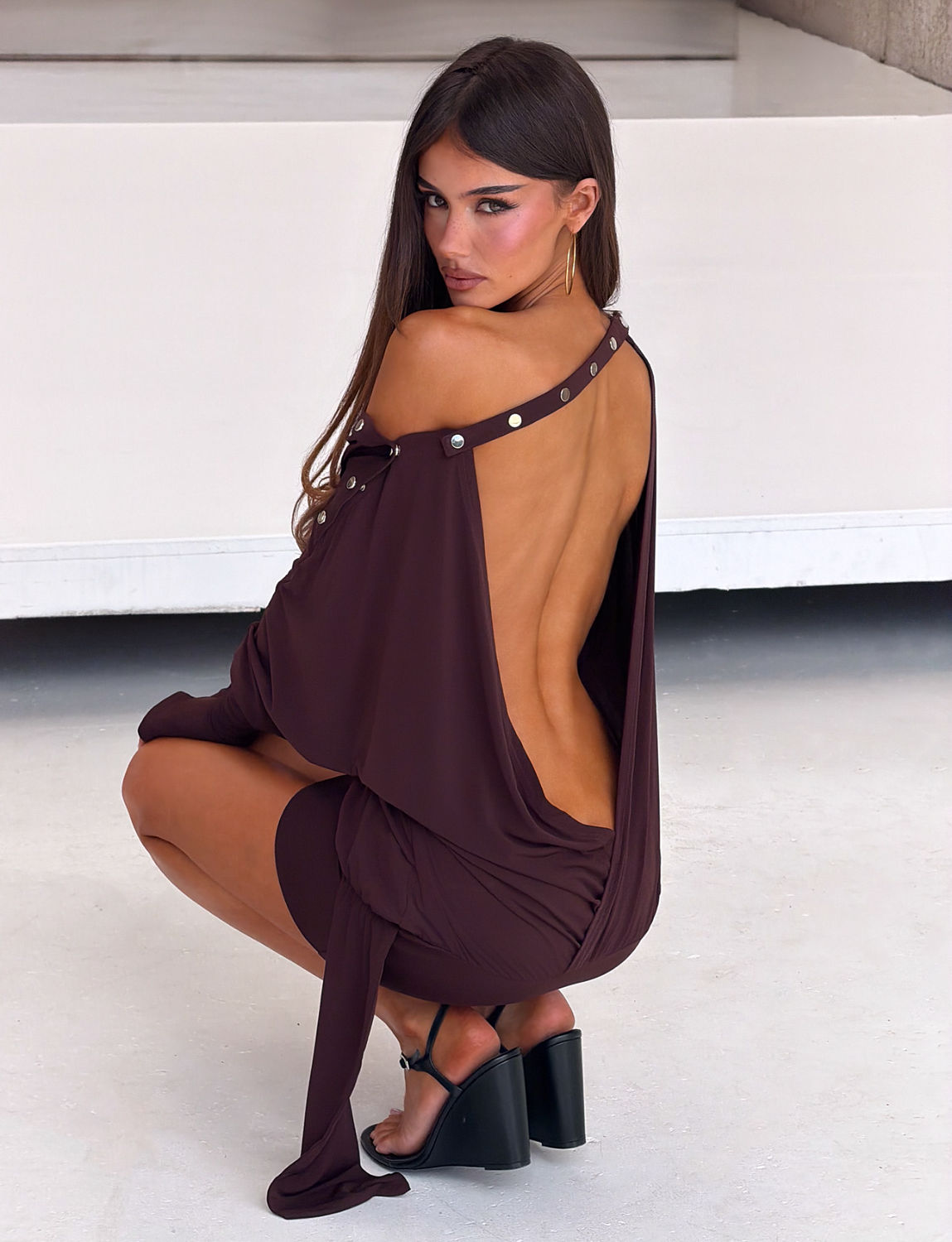 A woman with long brown hair squats while facing backward, wearing the TIGERMIST RAVENNA LONG SLEEVE MINI DRESS in Dark Chocolate with metal studs on the straps and black heeled sandals in a minimalist, light-colored setting.