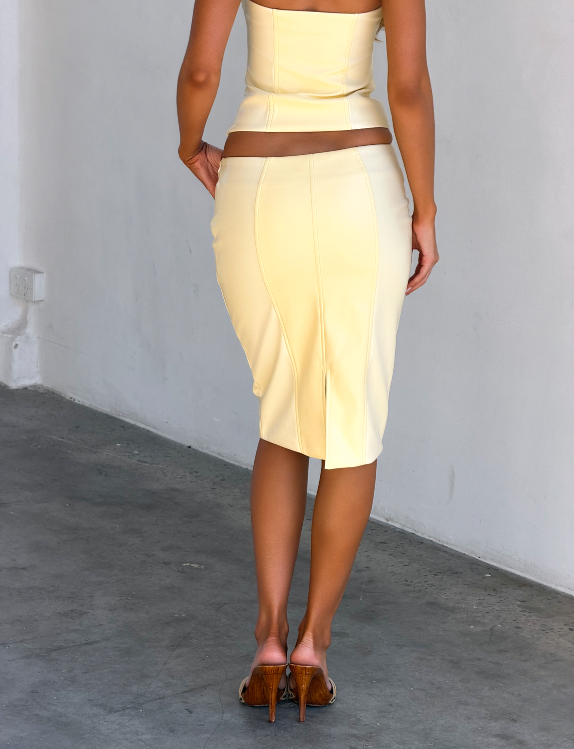 A woman stands indoors facing away, wearing the TIGERMIST EMERALD MIDI SKIRT - BUTTER YELLOW with a matching strapless top and high heels against a white wall on a concrete floor.