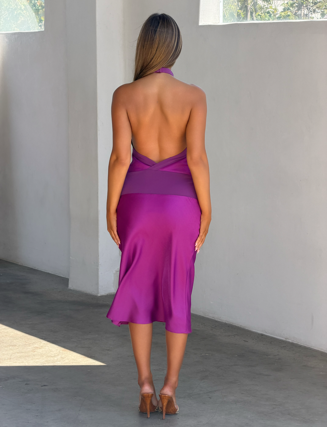 A woman stands indoors with her back to the camera, wearing the ZAYA MIDI DRESS - MULBERRY by TIGERMIST and clear heels. Sunlight streams through windows, casting shadows on the pale floor and wall.