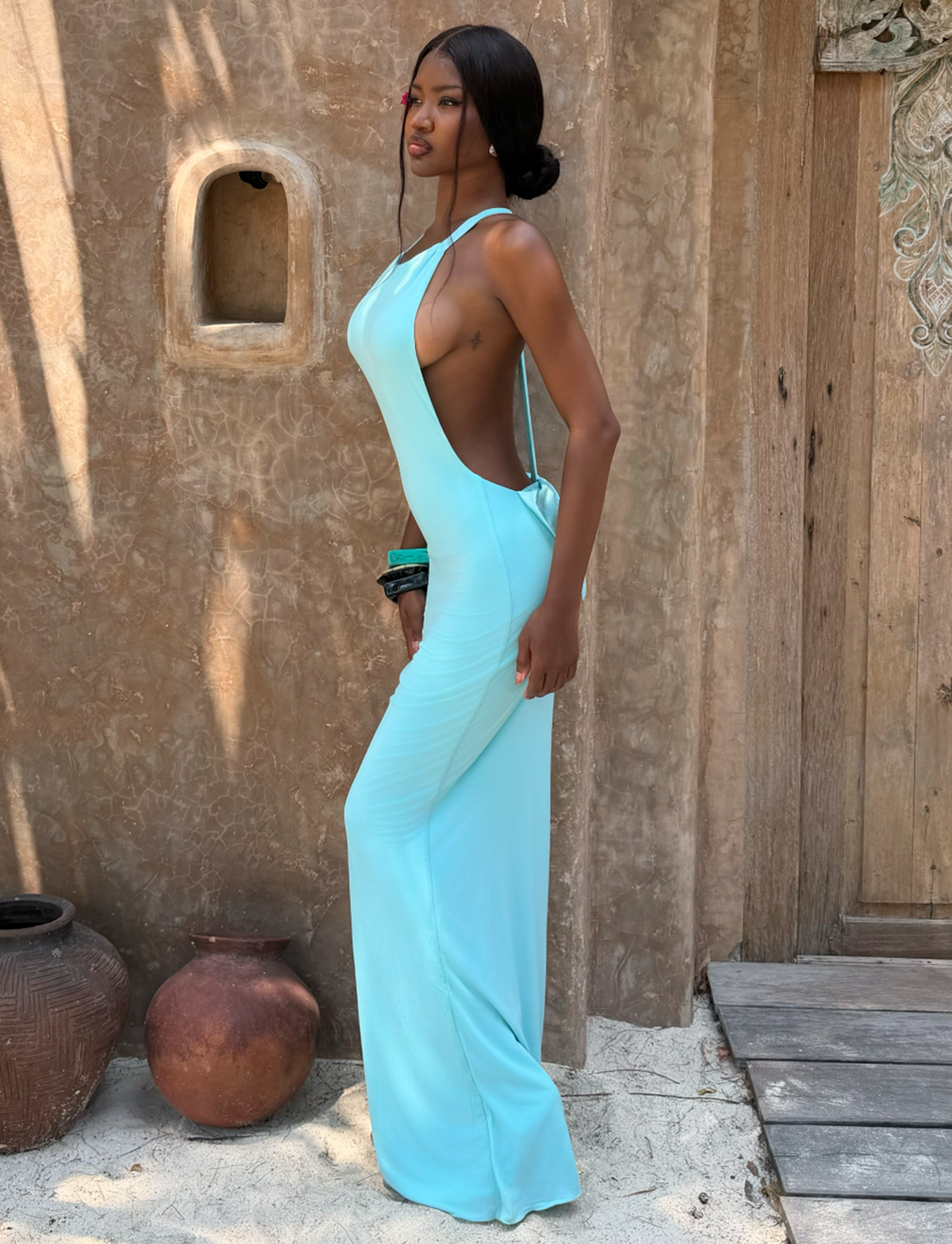 A woman wears the TIGERMIST RONI MAXI DRESS - AQUA, standing on sand by a rustic wall with clay pots and a wooden door, gazing to the left.