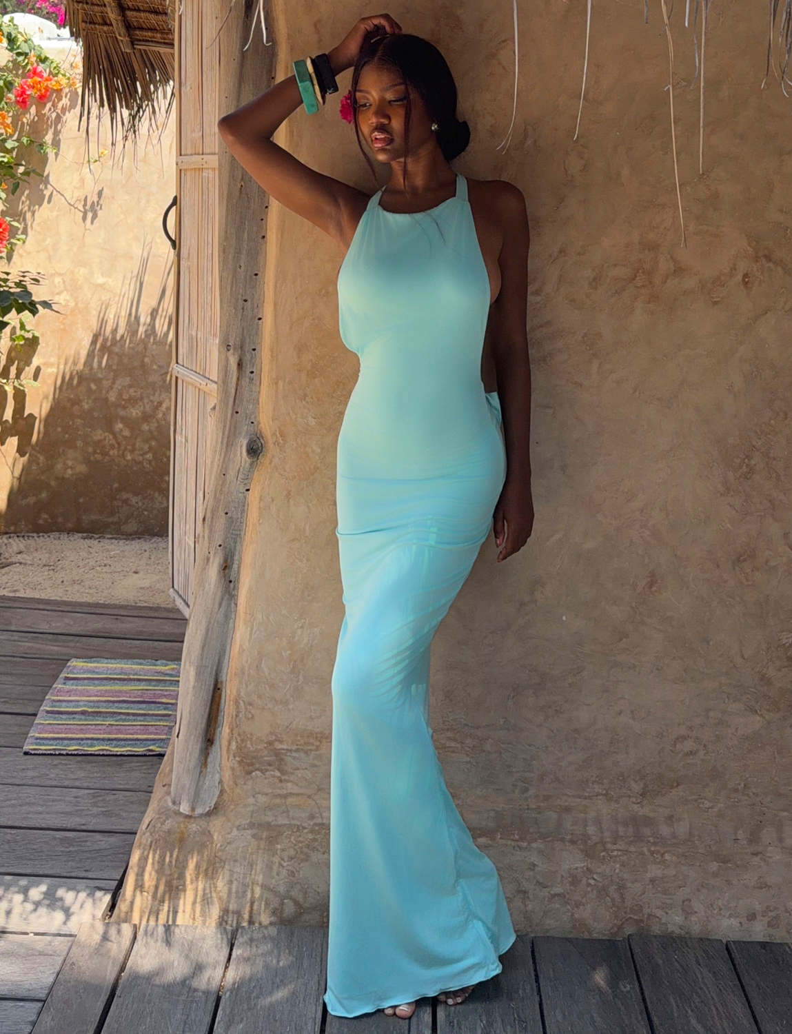 A woman in the RONI MAXI DRESS - AQUA by TIGERMIST poses by a rustic wall, one hand on her head, beneath a shaded structure on a sunny day.