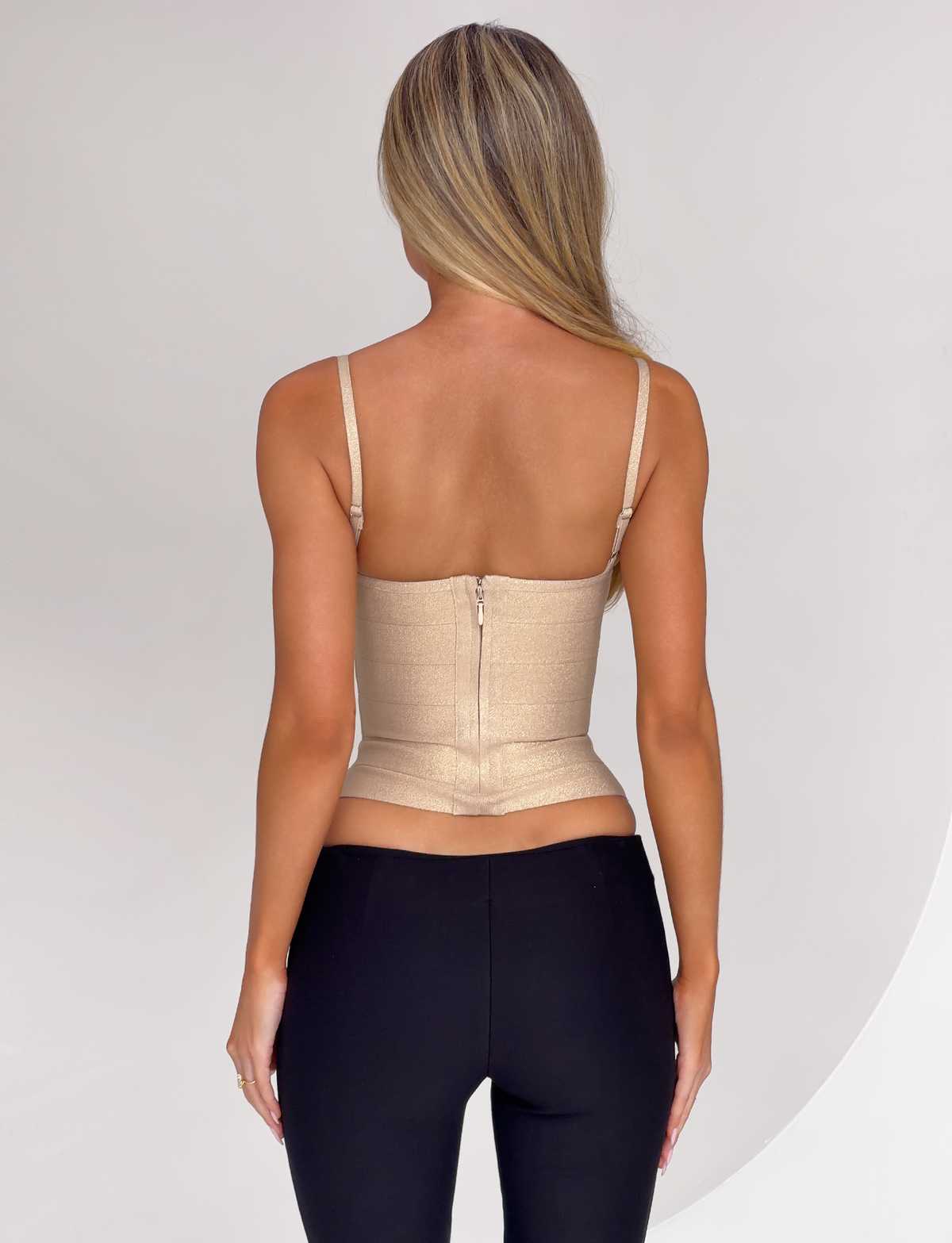 A sparkly gold MIDORI BANDAGE TOP - GOLD SPARKLE featuring a corset-style design, thin straps, and a center-back zipper, worn with black pants.