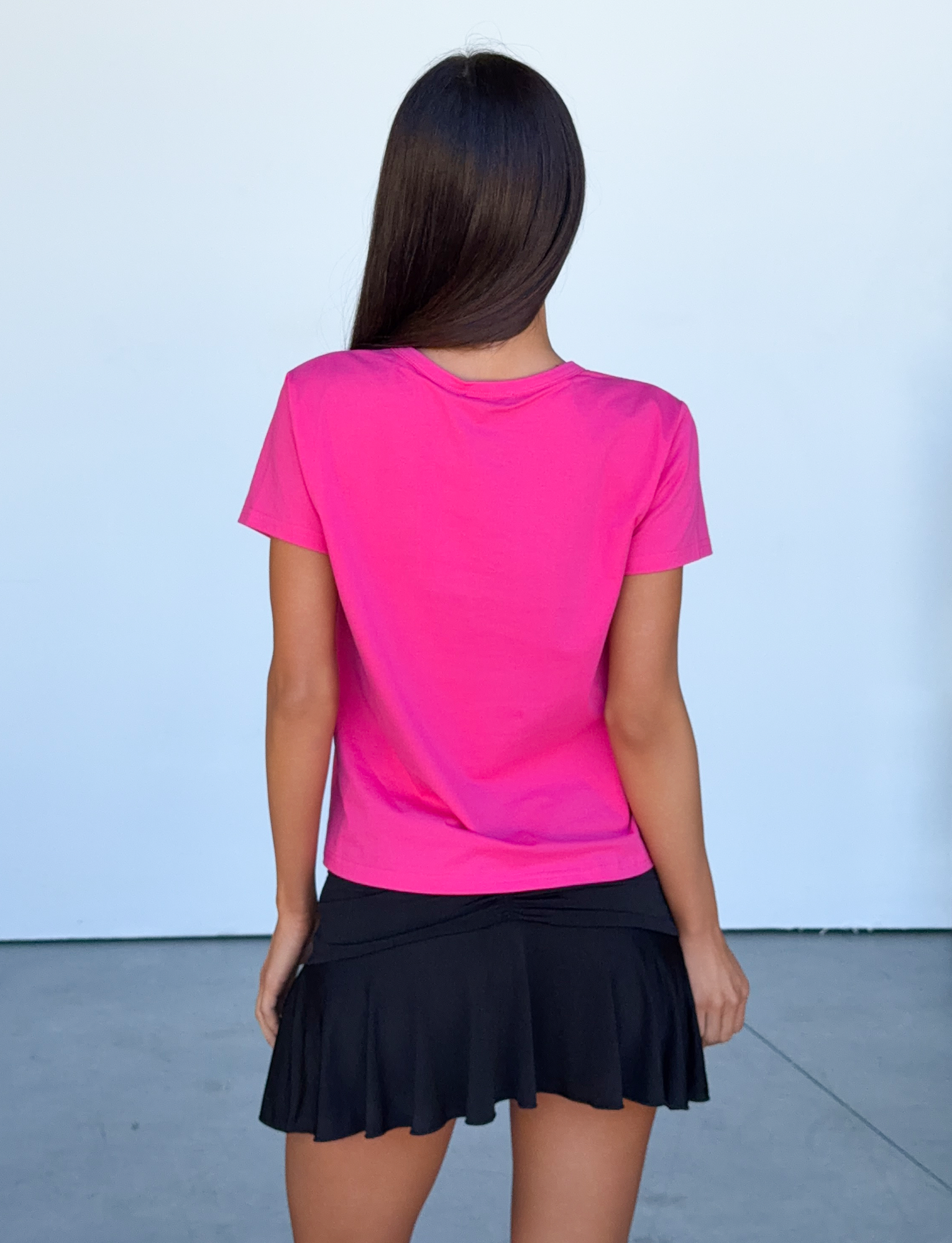 A woman with long brown hair stands facing away, wearing the TIGERMIST BELLA BABY TEE in hot pink and a short black skirt, against a plain light background.