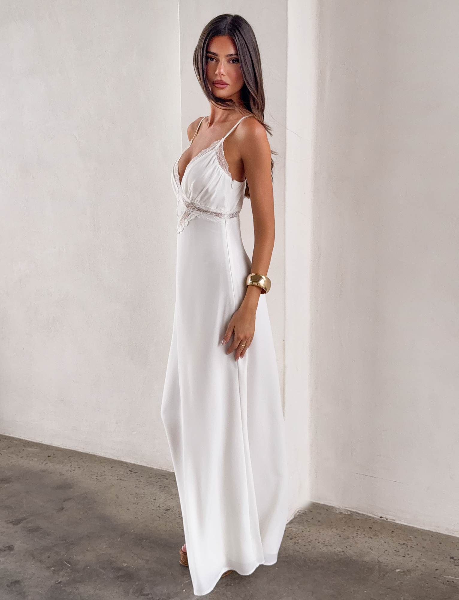 A woman models the TIGERMIST AISLYNN MAXI DRESS in white, featuring lace details. She stands against a plain light wall, wearing a gold bracelet and has long, straight brown hair.