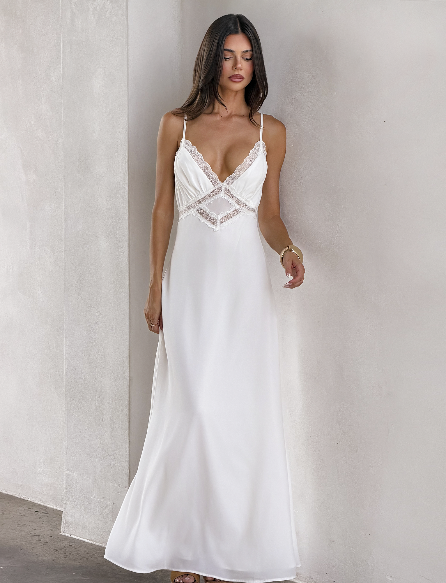 A woman with long dark hair wears the TIGERMIST AISLYNN MAXI DRESS - WHITE, a slip maxi dress with lace details and thin straps, standing against a light-colored wall.