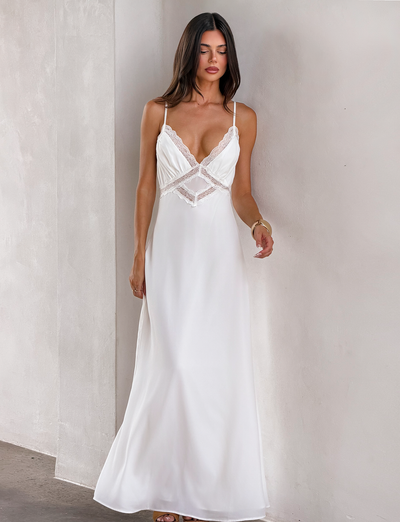 A woman with long dark hair wears the TIGERMIST AISLYNN MAXI DRESS - WHITE, a slip maxi dress with lace details and thin straps, standing against a light-colored wall.