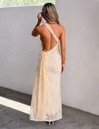 A woman with blonde hair stands indoors, facing away, wearing the TIGERMIST CHARLYSE ONE SHOULDER MAXI DRESS in butter yellow. She raises a hand near her head and pairs the look with white heels.