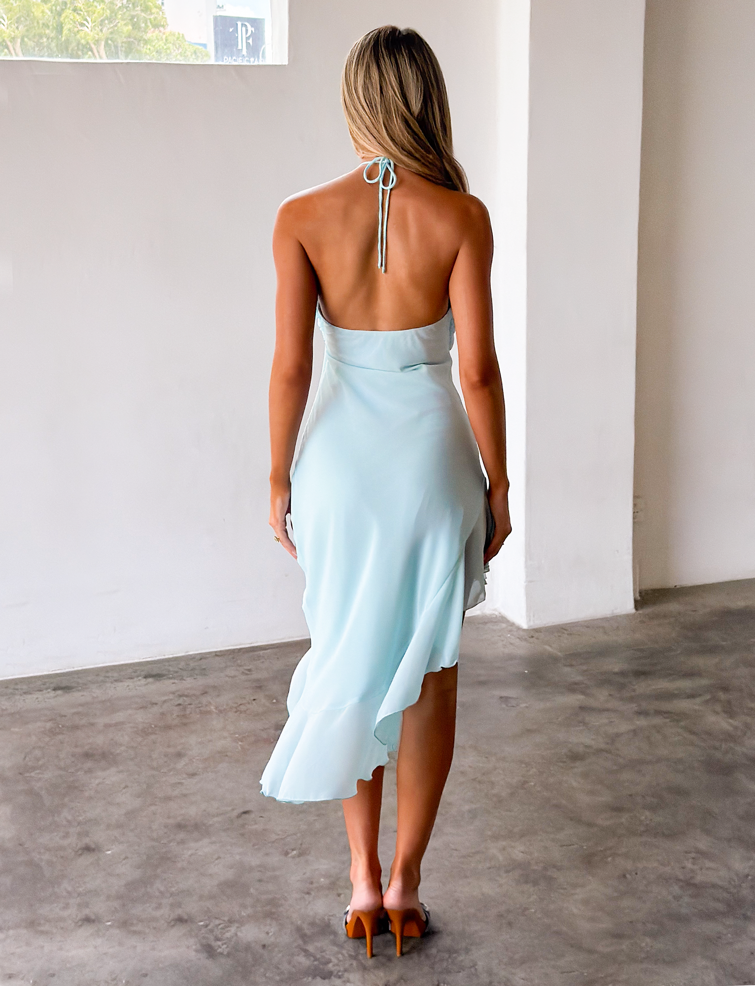 A woman with long blonde hair stands indoors on a concrete floor, facing away. She wears the TIGERMIST ELARINA MIDI DRESS in Mermaid Blue and brown heeled sandals.