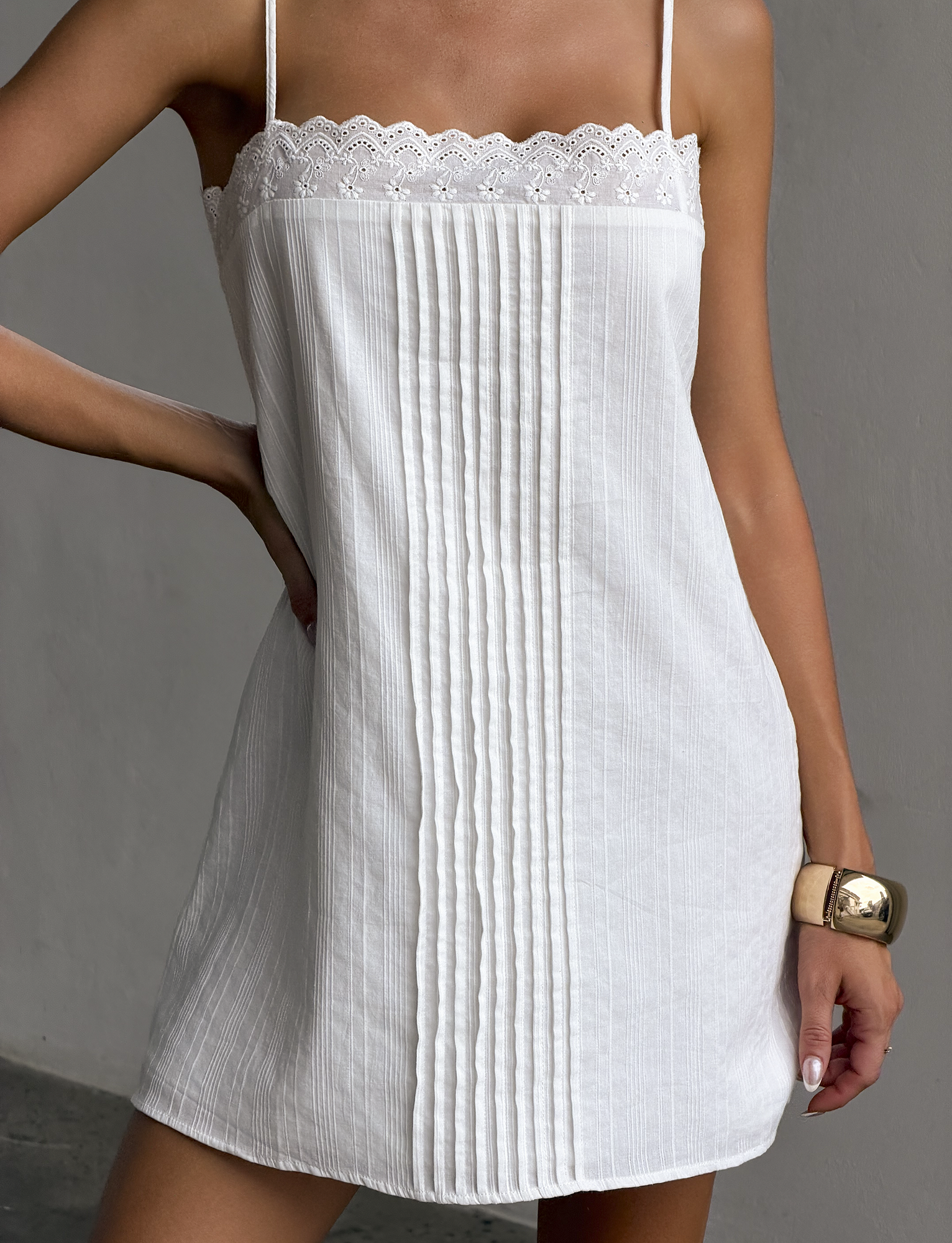 A woman models the TIGERMIST BRYNLEE MINI DRESS in white, a sleeveless style with lace trim, vertical pleats, and adjustable straps. She accessorizes with a chunky gold bracelet against a plain gray background.
