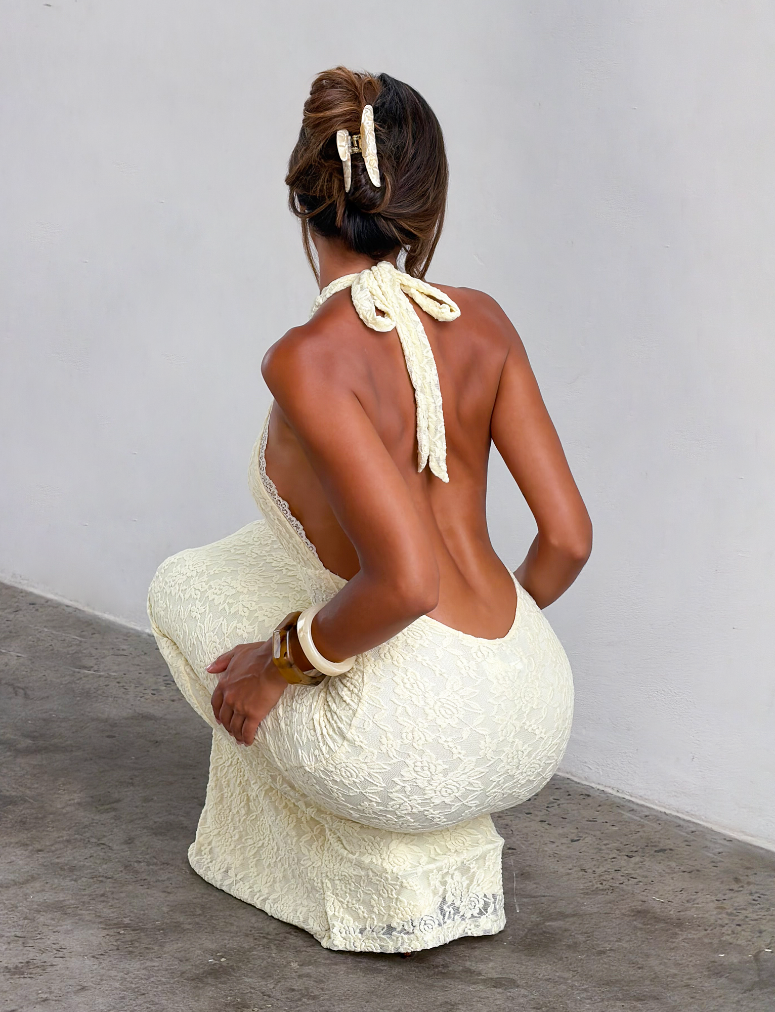 A woman with tan skin faces away, wearing the TIGERMIST MAVEN LACE MAXI DRESS in pastel yellow. The backless, halter style shows off her updo, matching pastel hair accessory, and bangles as she poses by a plain wall.